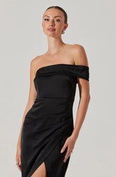 A woman is wearing a strapless black dress with a fitted bodice and a slightly off-the-shoulder draped neckline. The dress has a smooth, shiny texture with subtle gathers and a high slit on one side. She has her hair pulled back, and she is accessorized with earrings and rings.